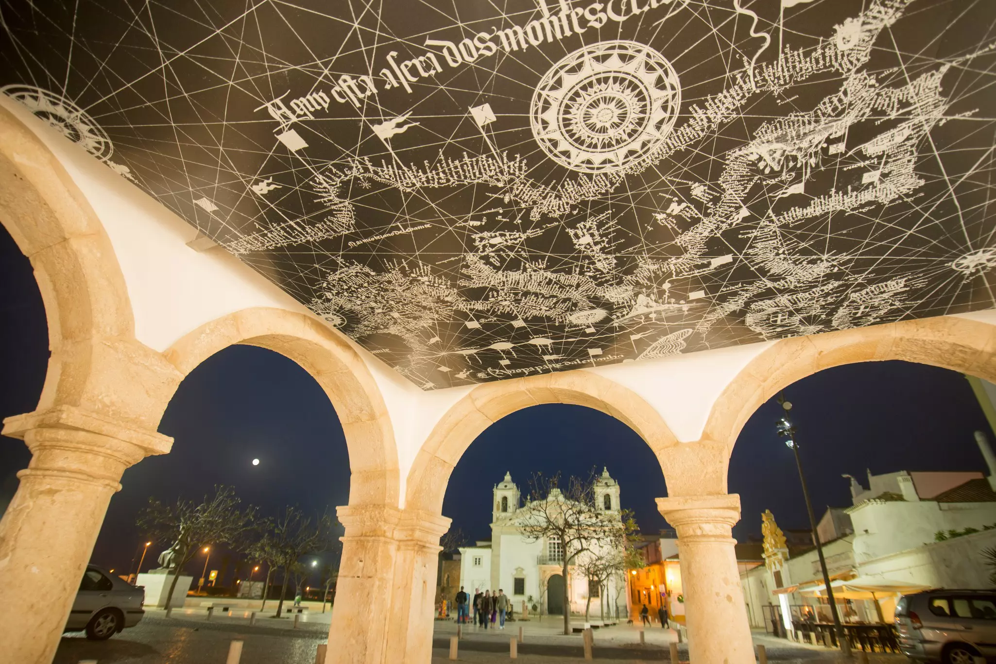 The archways of a pavilion in Lagos, Portugal, is pictured at night. A ceiling mural depicts a map, while a church is visible in the square beyond.