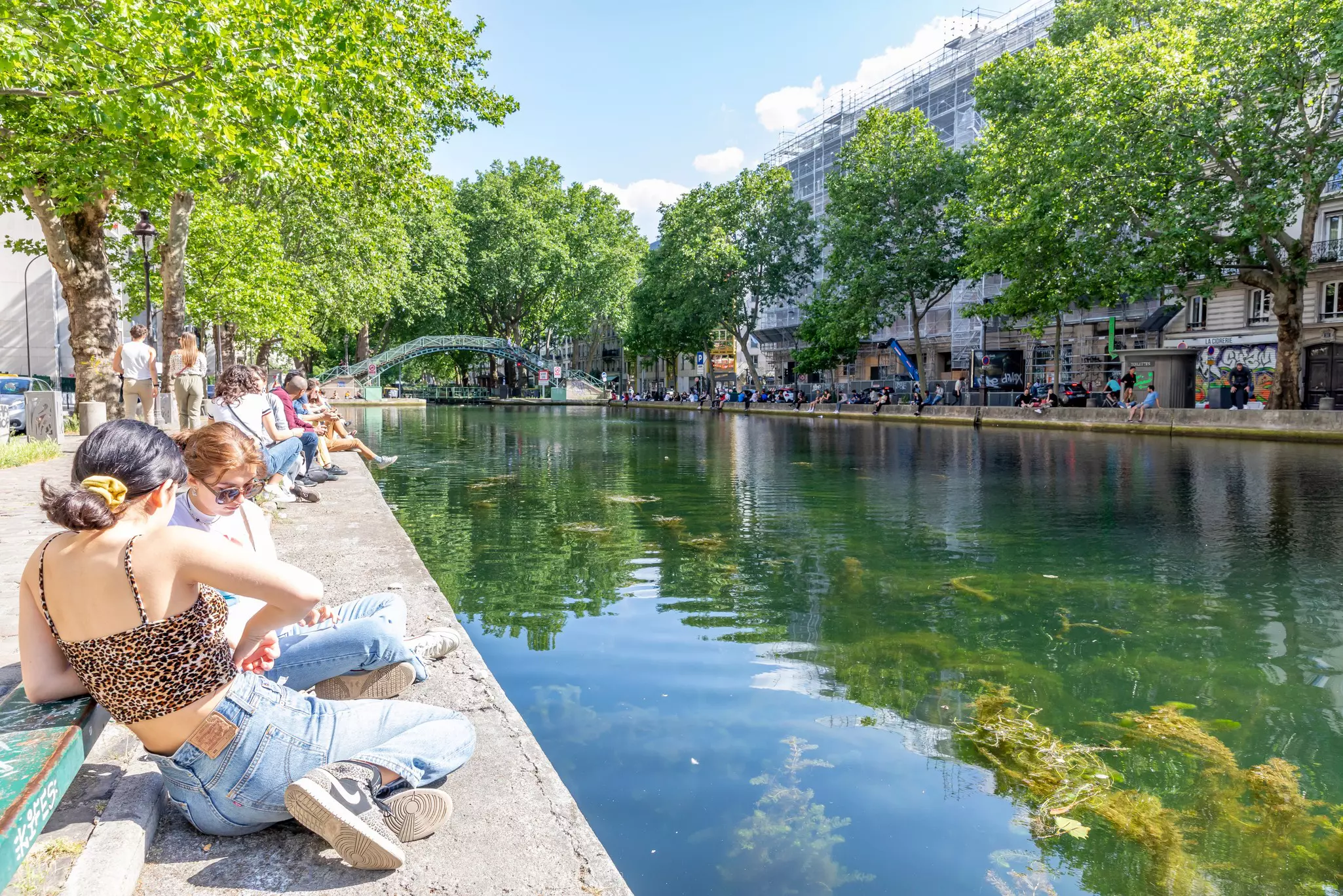 People sit in groups of friends beside a green tree-lined canal on a bright sunny day.