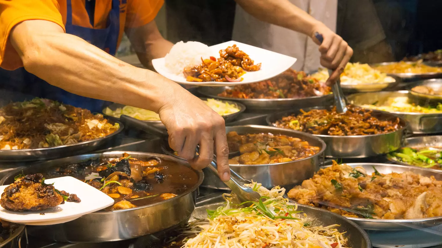 Two men with tongs select ingredients from among numerous bowls filled with food at a street food stall.