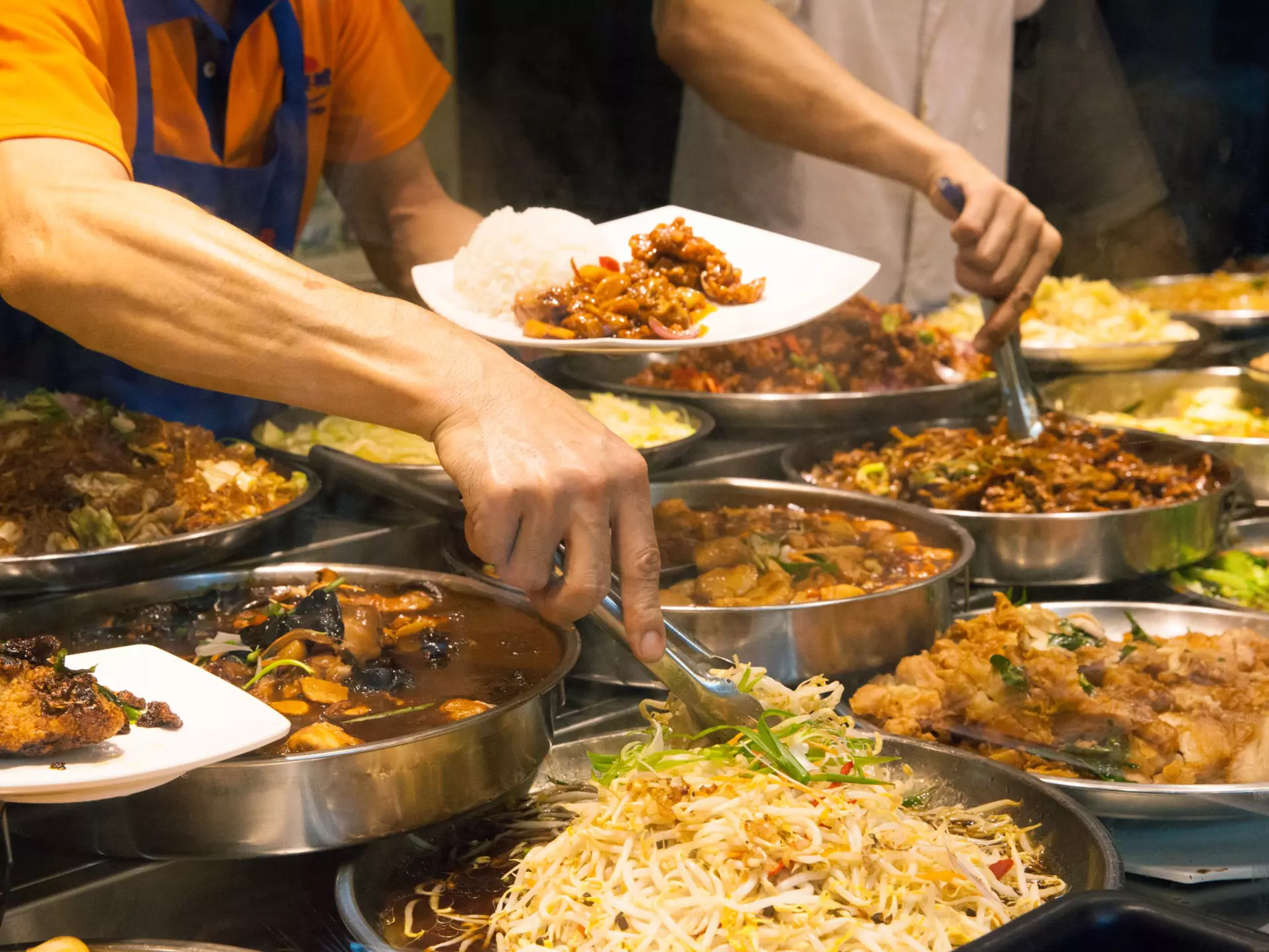 Two men with tongs select ingredients from among numerous bowls filled with food at a street food stall.