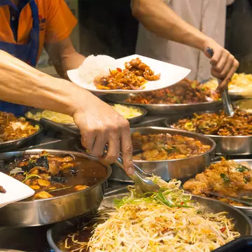 Two men with tongs select ingredients from among numerous bowls filled with food at a street food stall.