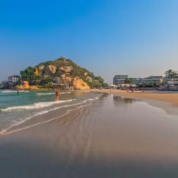 Khao Takiab headland in Hua Hin with a golden standing Buddha on its cliff, above a sandy beach with waves and hotels