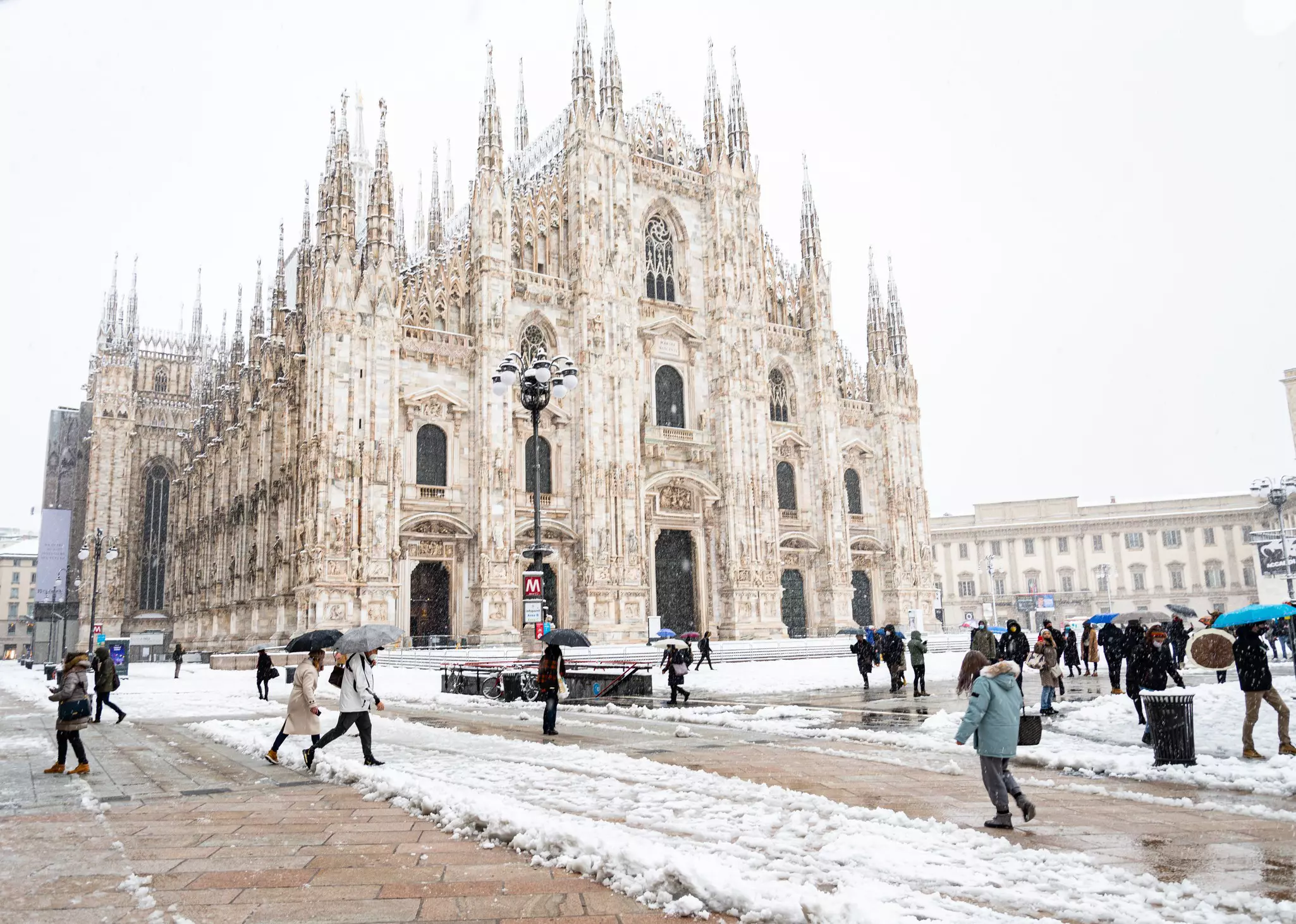 People cross a square on a snowy day, past a magnificent Gothic church. Piles of snow are seen on the street.
