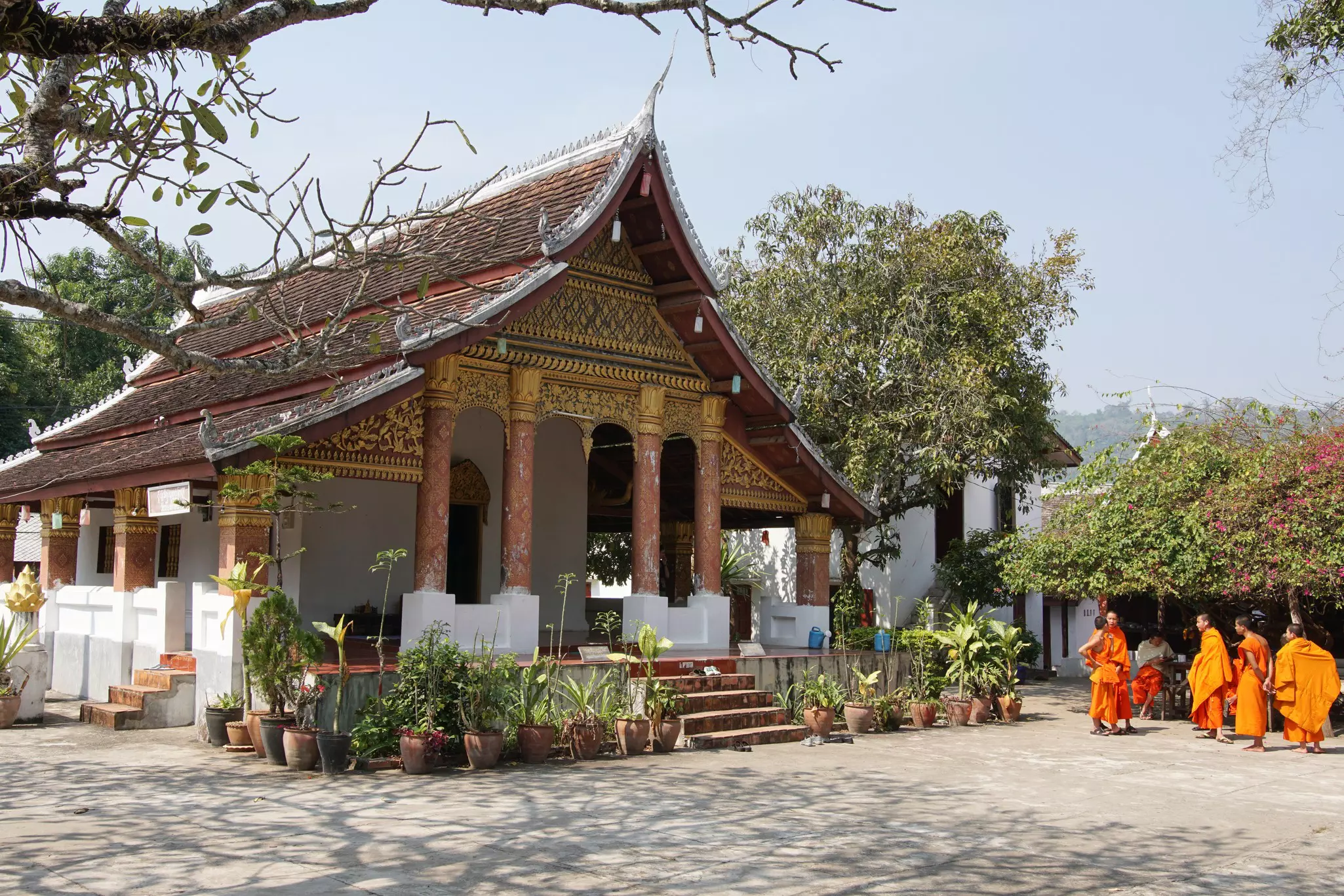 Buddhist monks dressed in orange robes stand in front of a temple with a brown-shingled roof, four pillars and an ornate gold facade