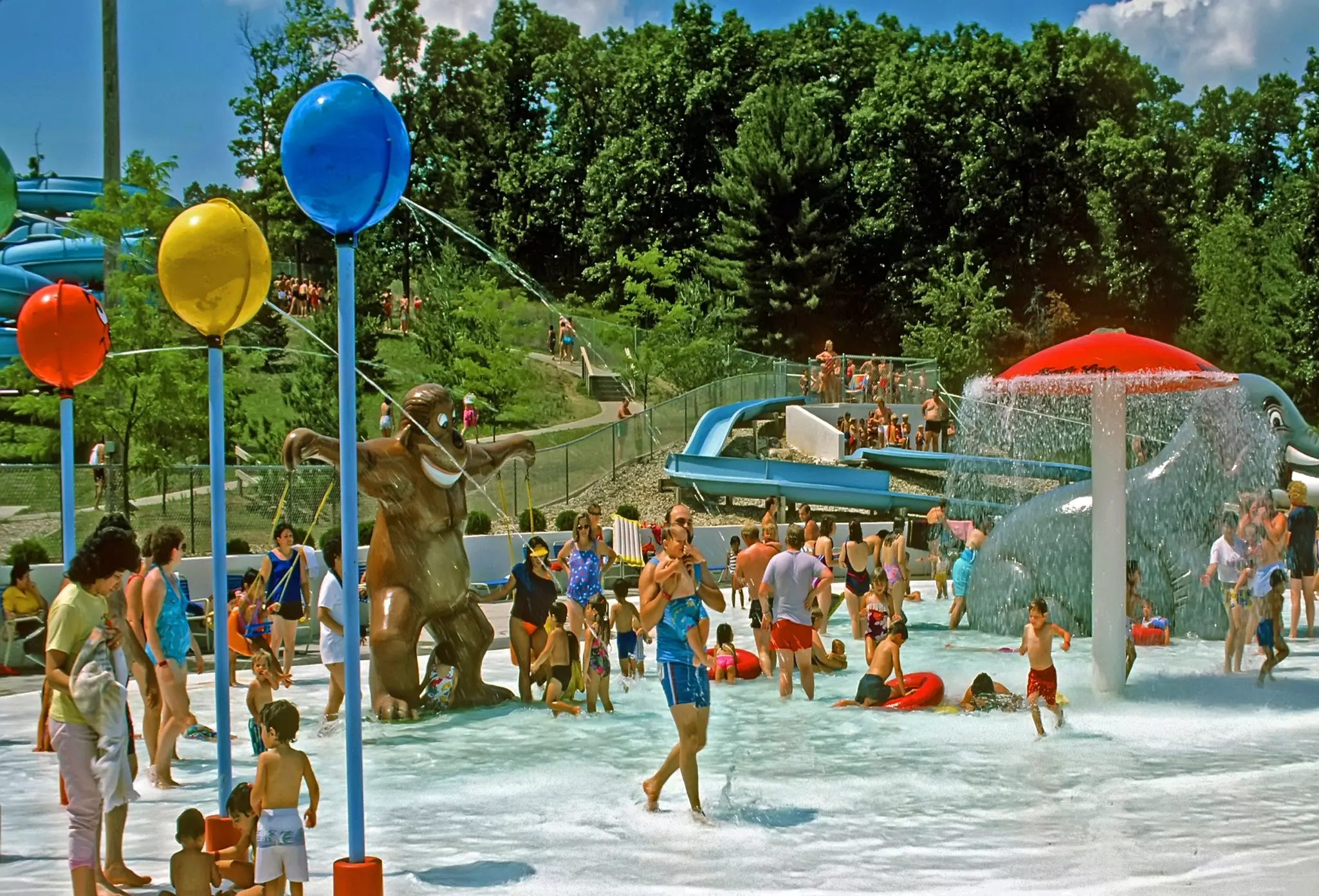 People of various ages are pictured at an outdoor water park with pools, fountains and other features.