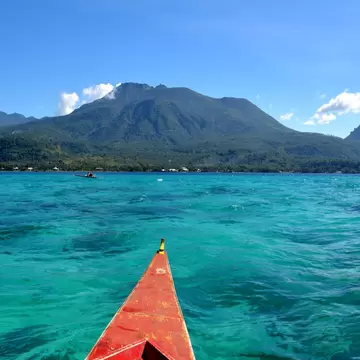 Red wooden boat bow pointing across bright turquoise water toward green volcanic mountains under a blue sky.