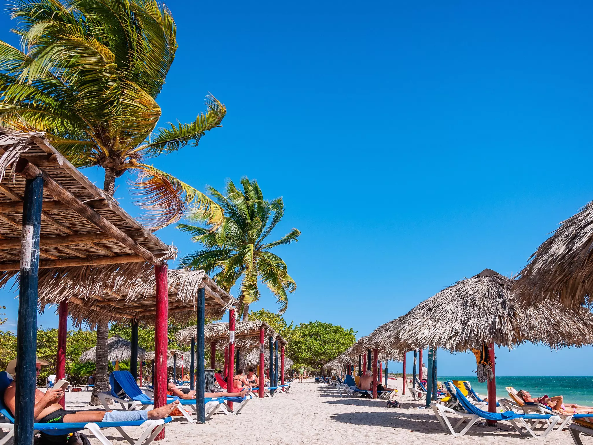 People sunbathing on lounge chairs under palm trees and parasols at the Playa Ancon at the Caribbean seaside near the city of Trinidad, Cuba.