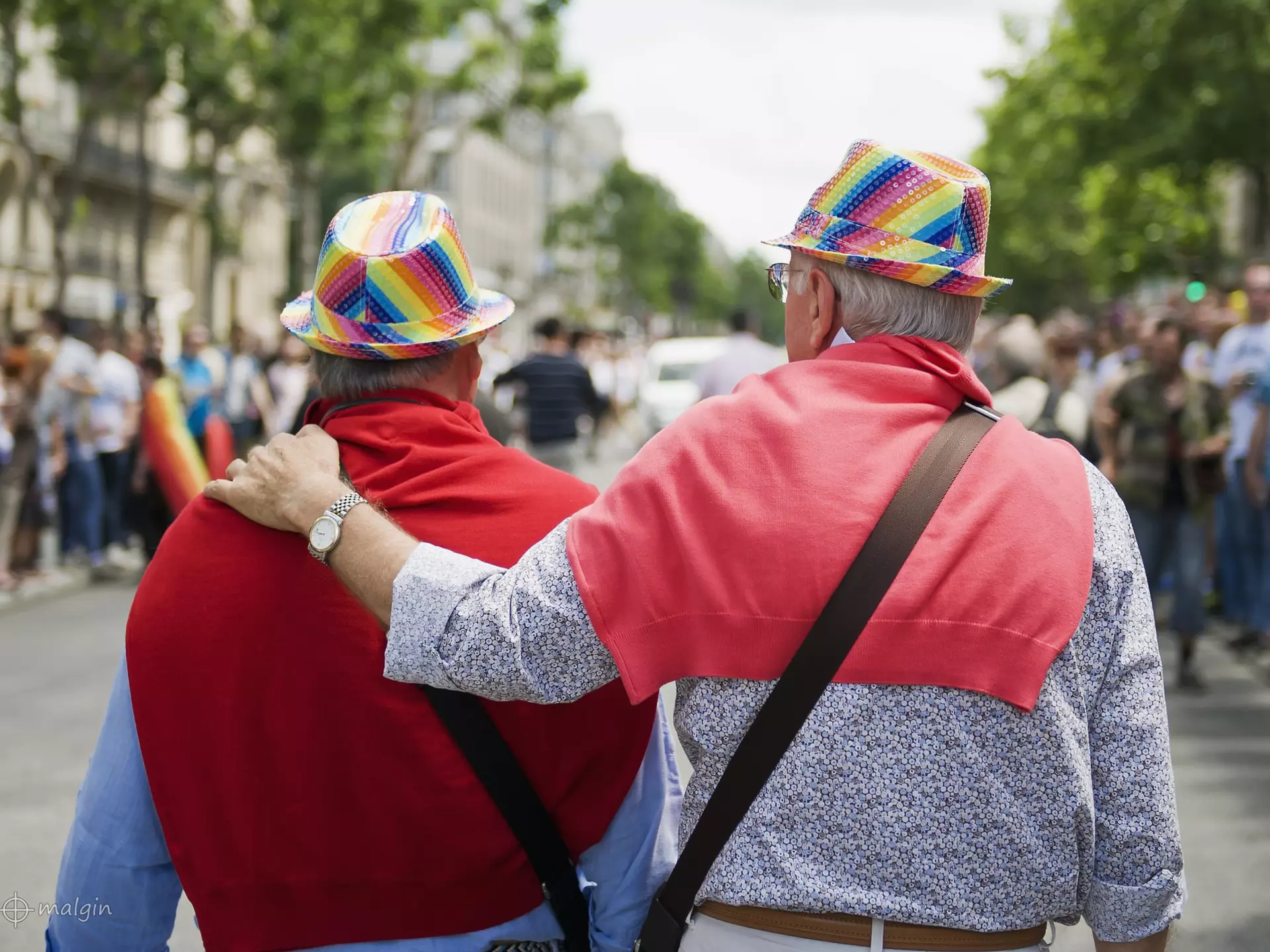 Two men viewed from behind. They're walking along together, one with his arm around the other. Both have red jumpers slung over their shoulders and are wearing rainbow hats