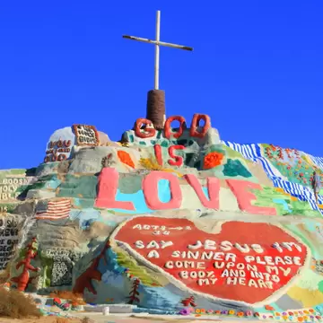 Hill of bright painted adobe topped by a white cross, with the words God Is Love and a large red heart, under blue sky