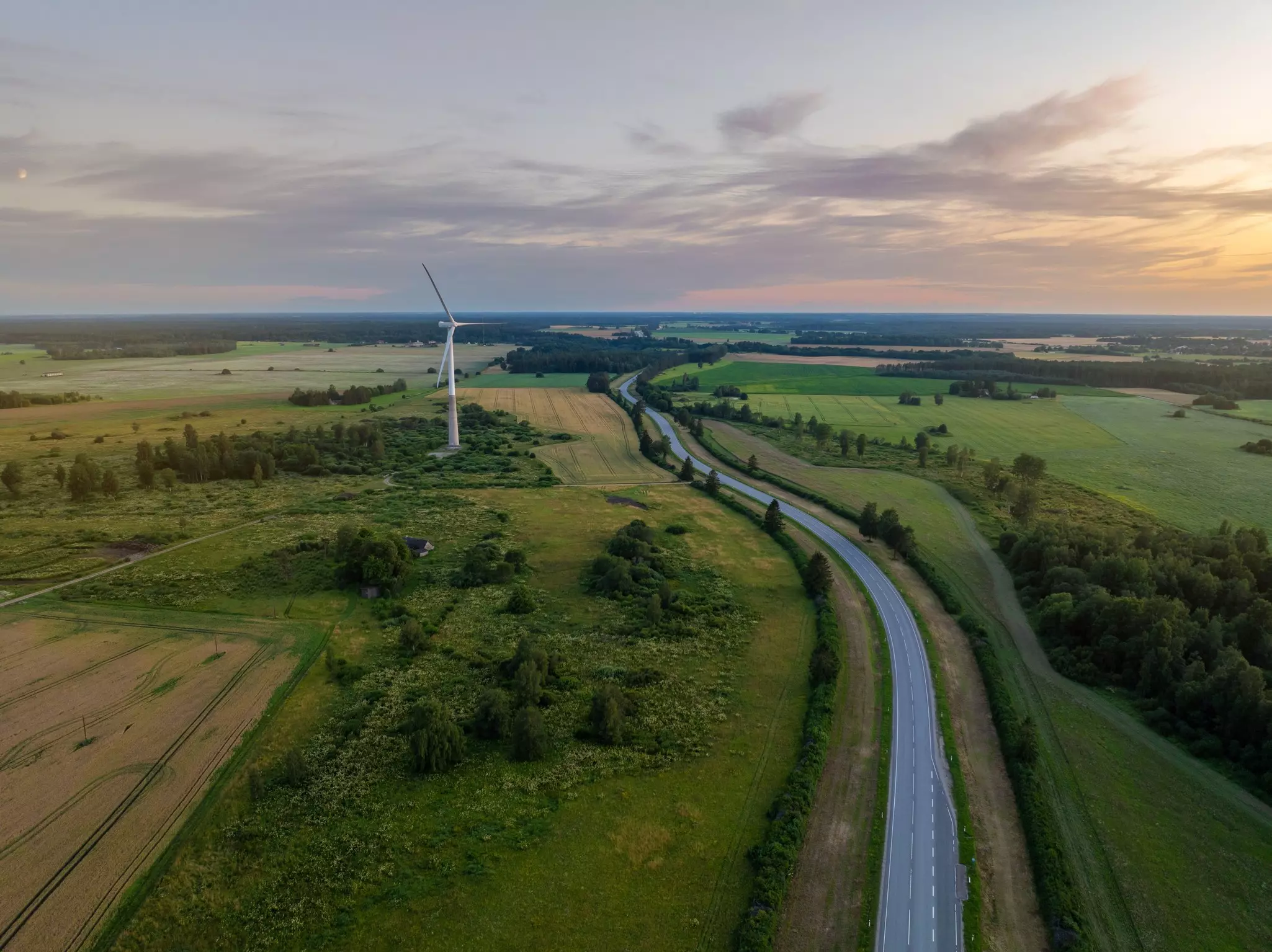 Aerial view of two-lane highway through green fields at sunset, with a modern windmill to the left.