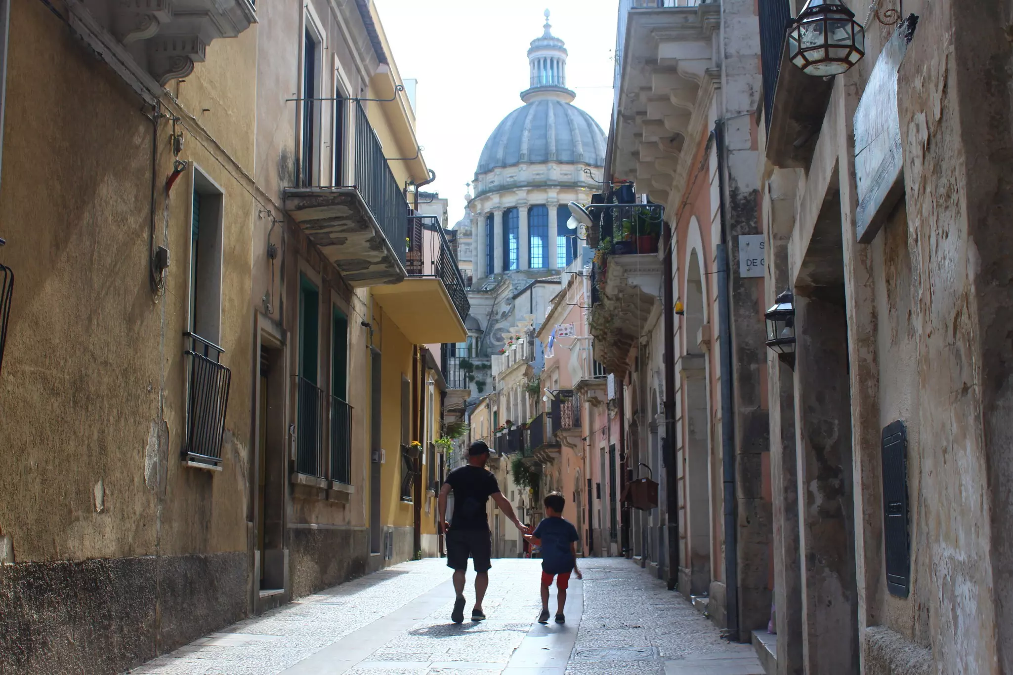 A man and a boy are seen walking from behind down a narrow pedestrianized street in a historic city. The dome of a church is visible in the distance.