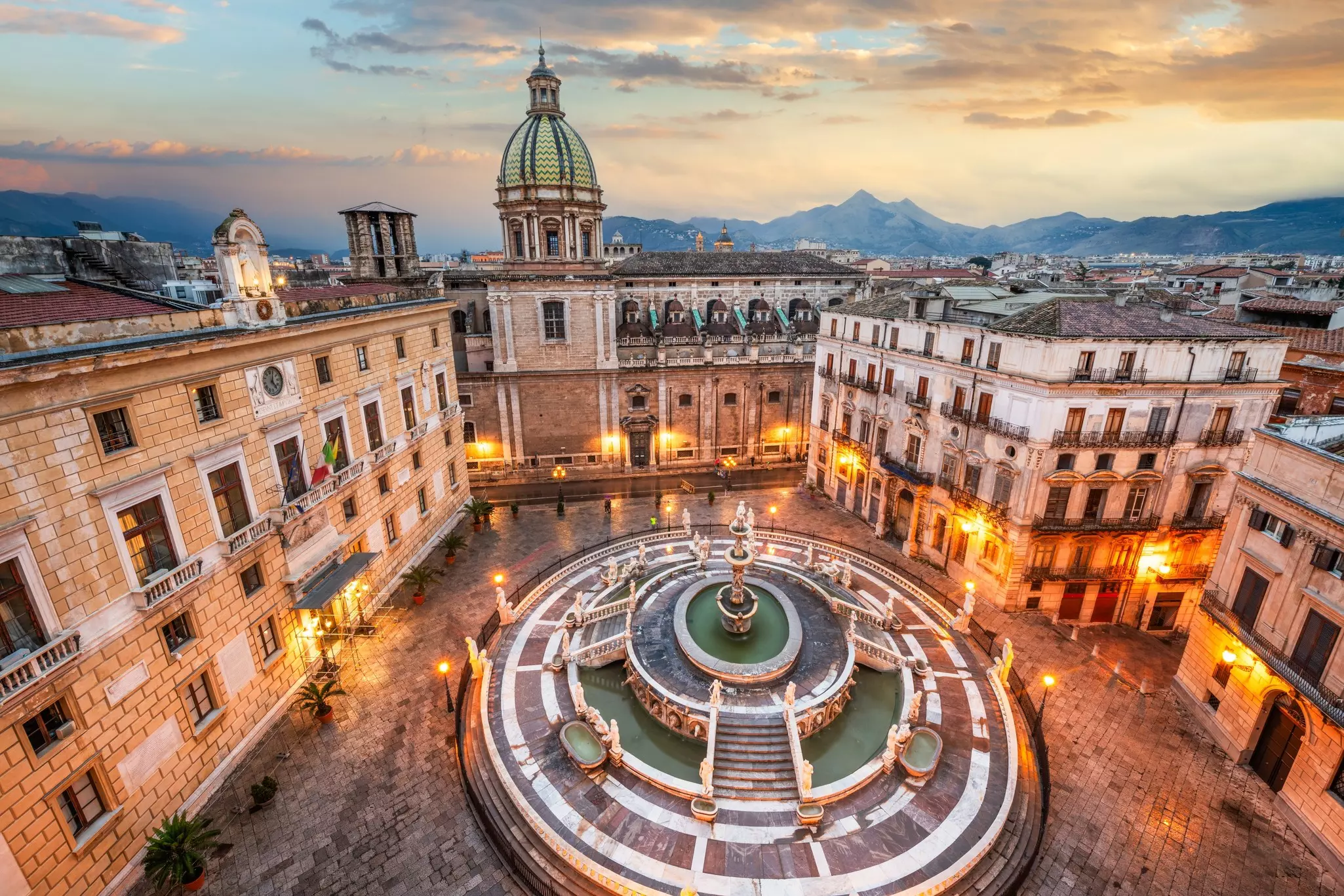 A large ornate fountain at the center of a city square overlooked by a domed church at dusk, Piazza Pretoria in Palermo, Sicily.