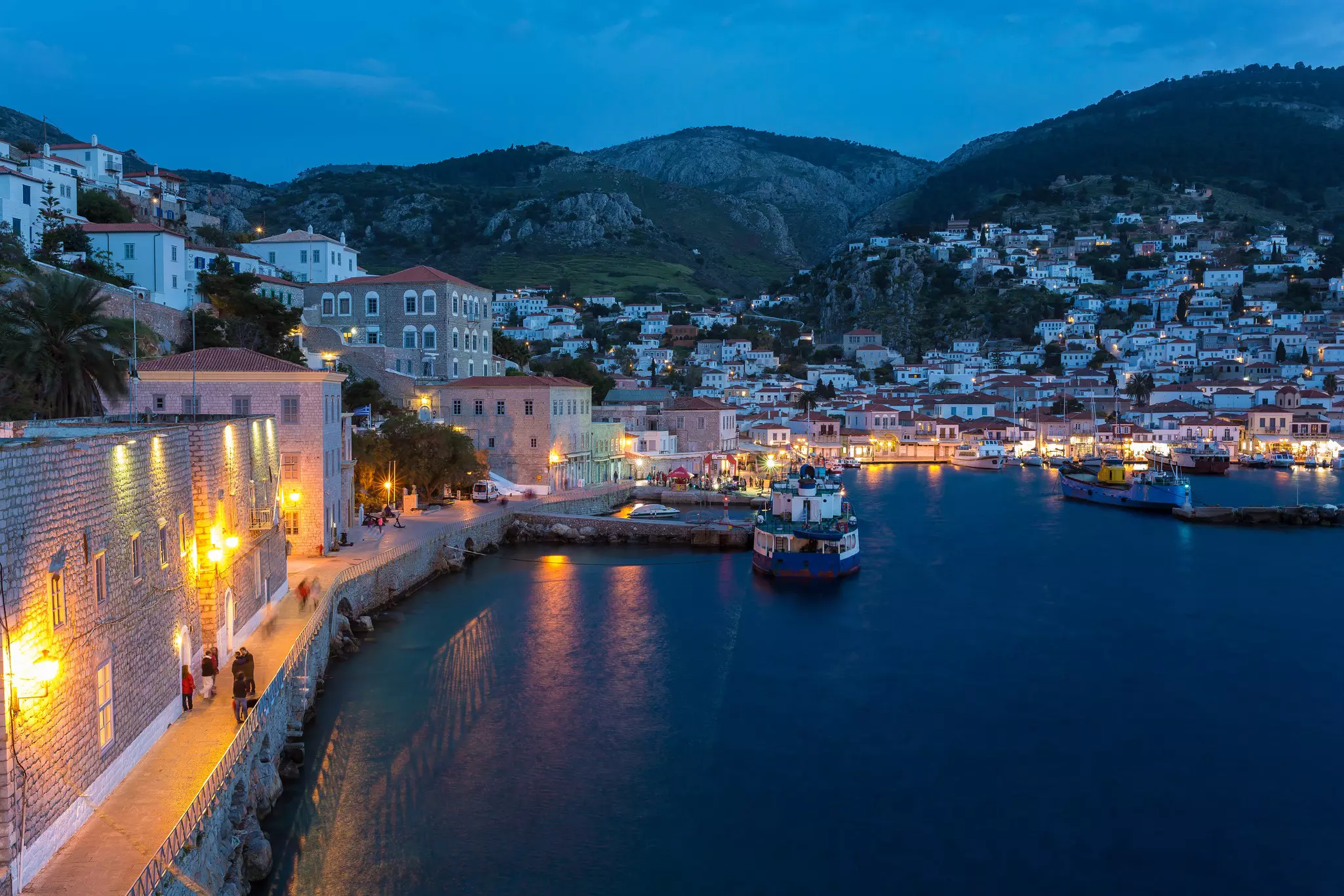 A wide view of a harbor with lights illuminating the promenade. White houses are seen on a hillside leading up from the water.