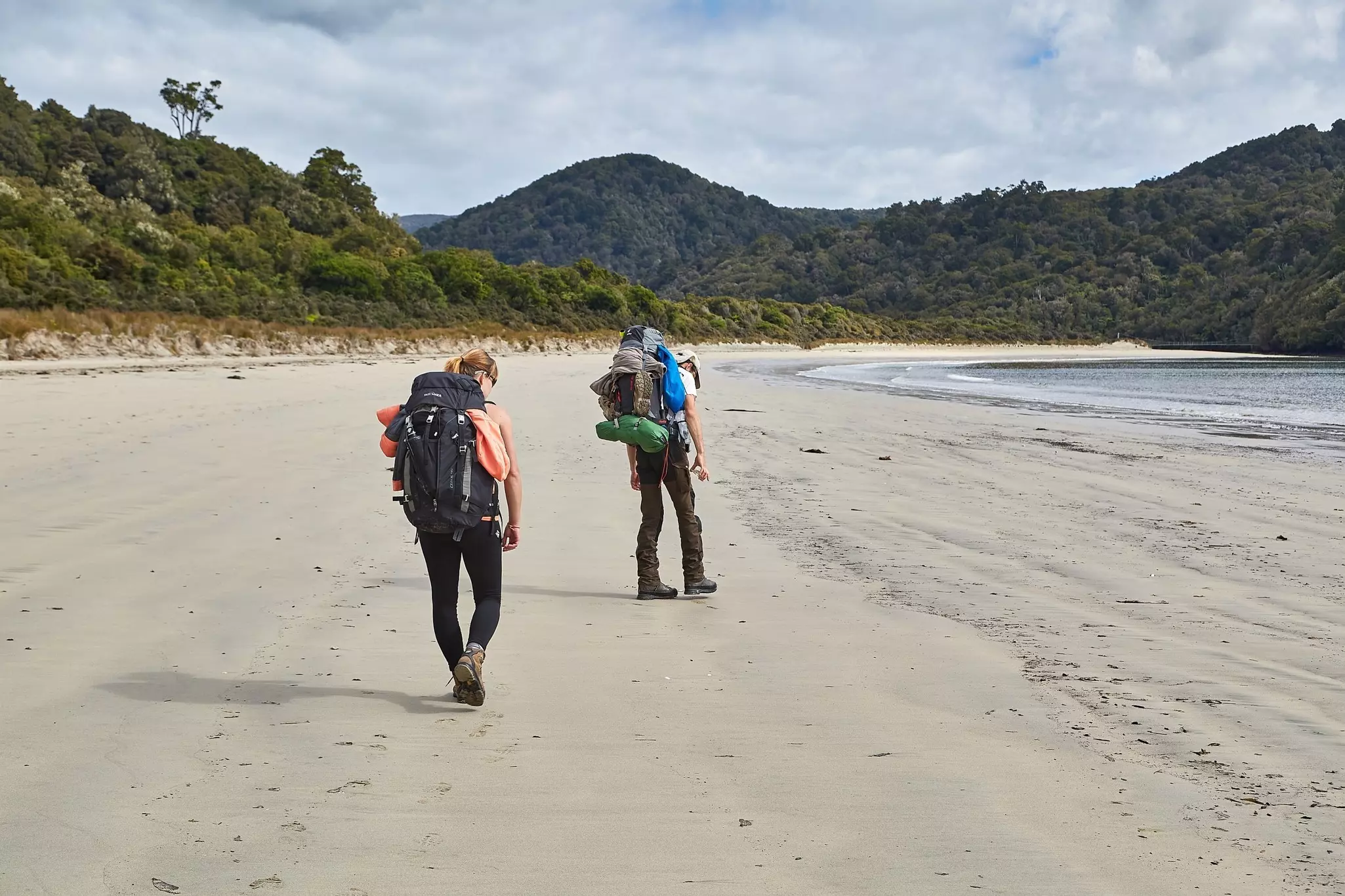 Man and woman with large backpacks on a wide sandy beach with forested hills in the distance.