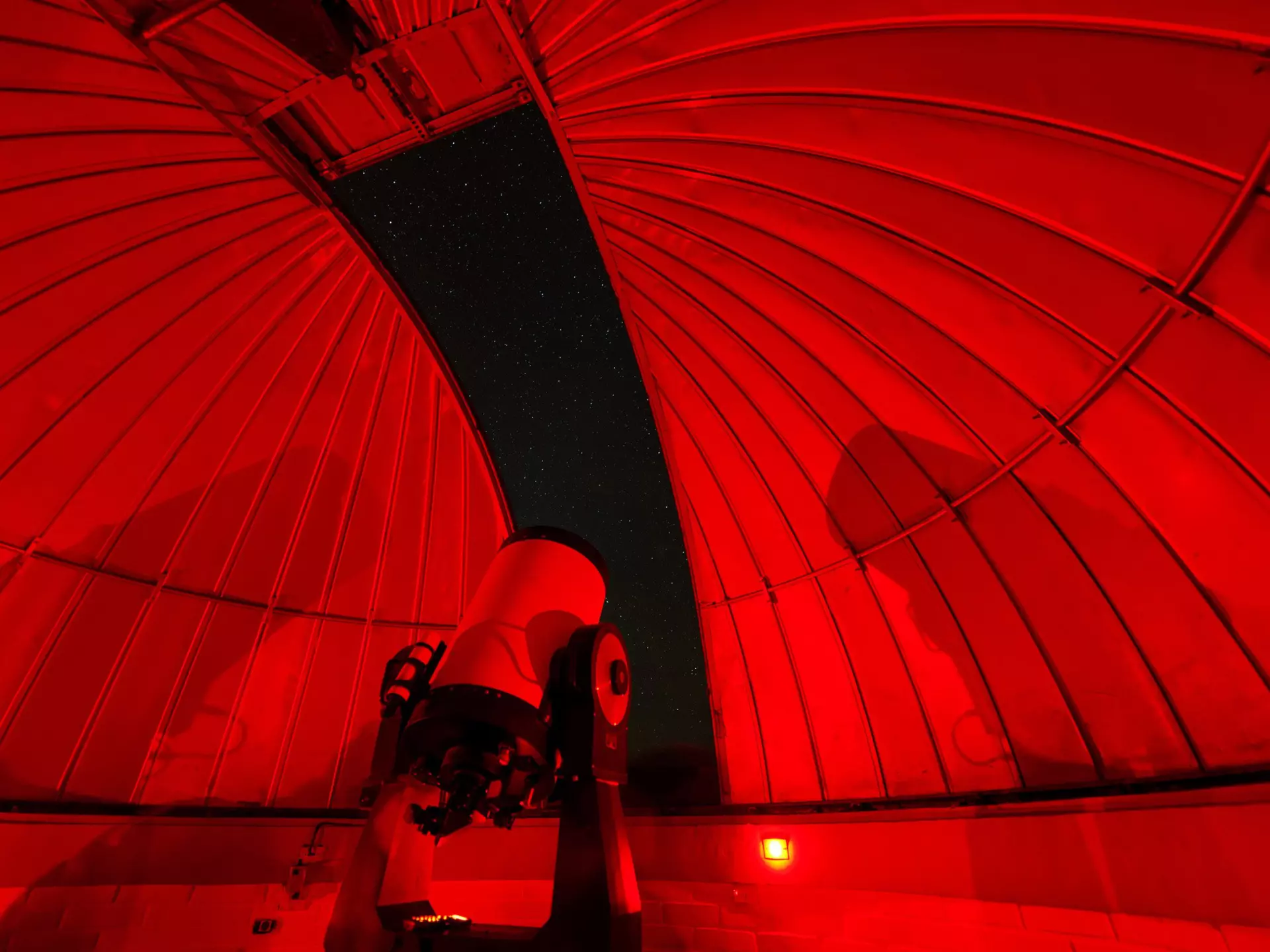 An observatory at night, lit with dim red light. A large telescope stands in the center of the observatory pointing towards the sky