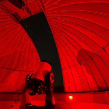 An observatory at night, lit with dim red light. A large telescope stands in the center of the observatory pointing towards the sky