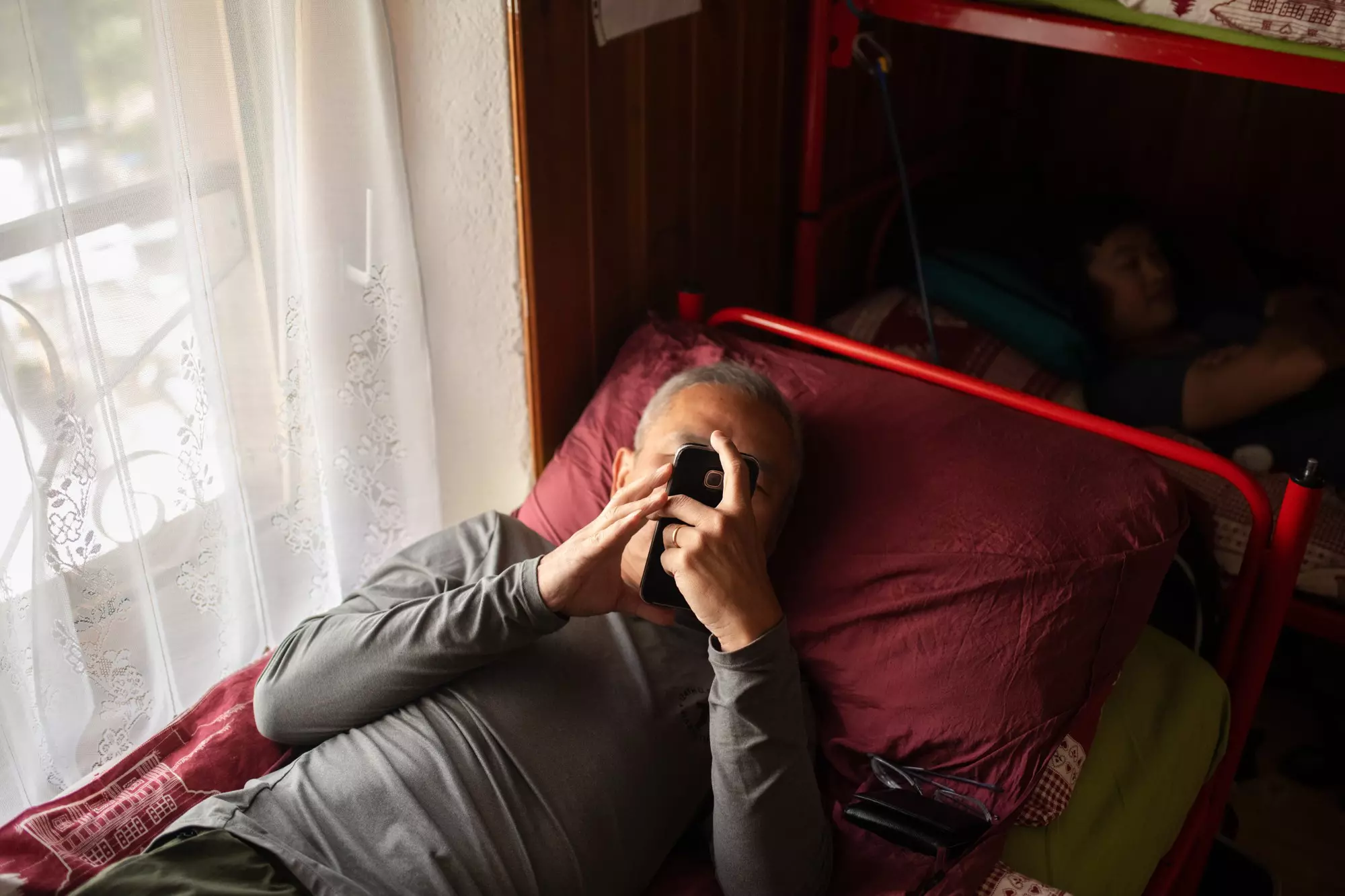 A man reclines in a bunk bed while looking at a smartphone.