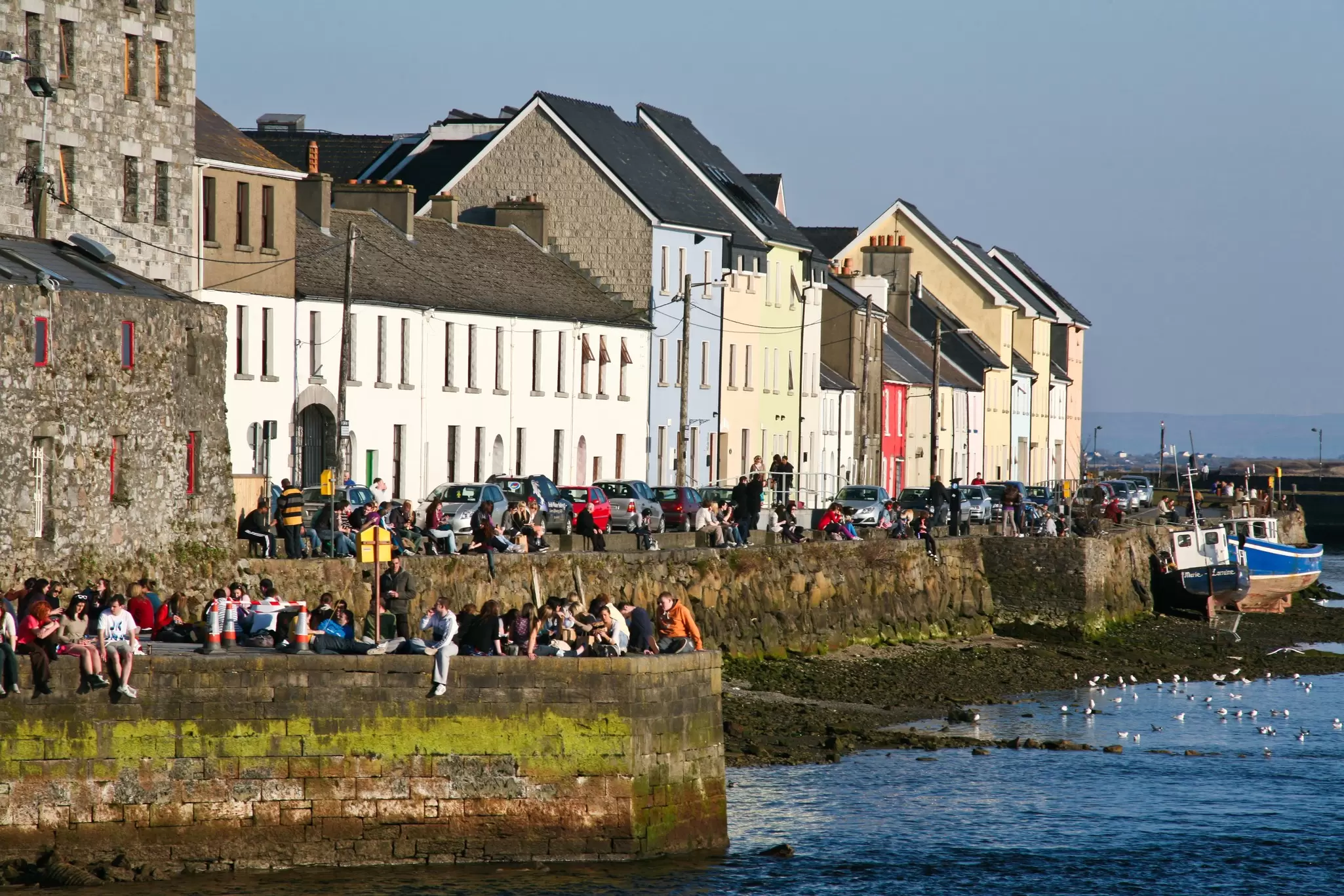 People sit on a sea wall in sunshine, with low buildings behind them.