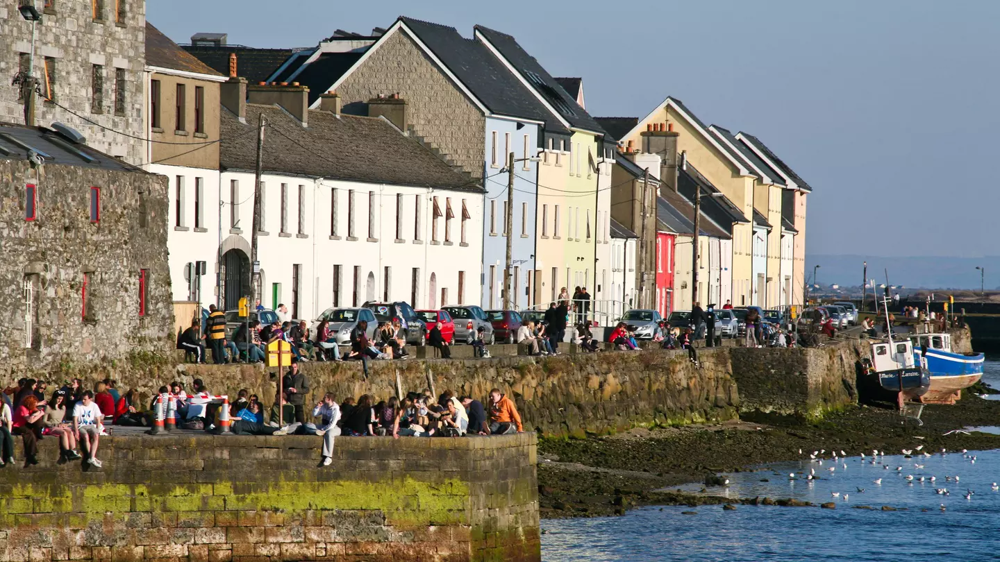 People sit on a sea wall in sunshine, with low buildings behind them.