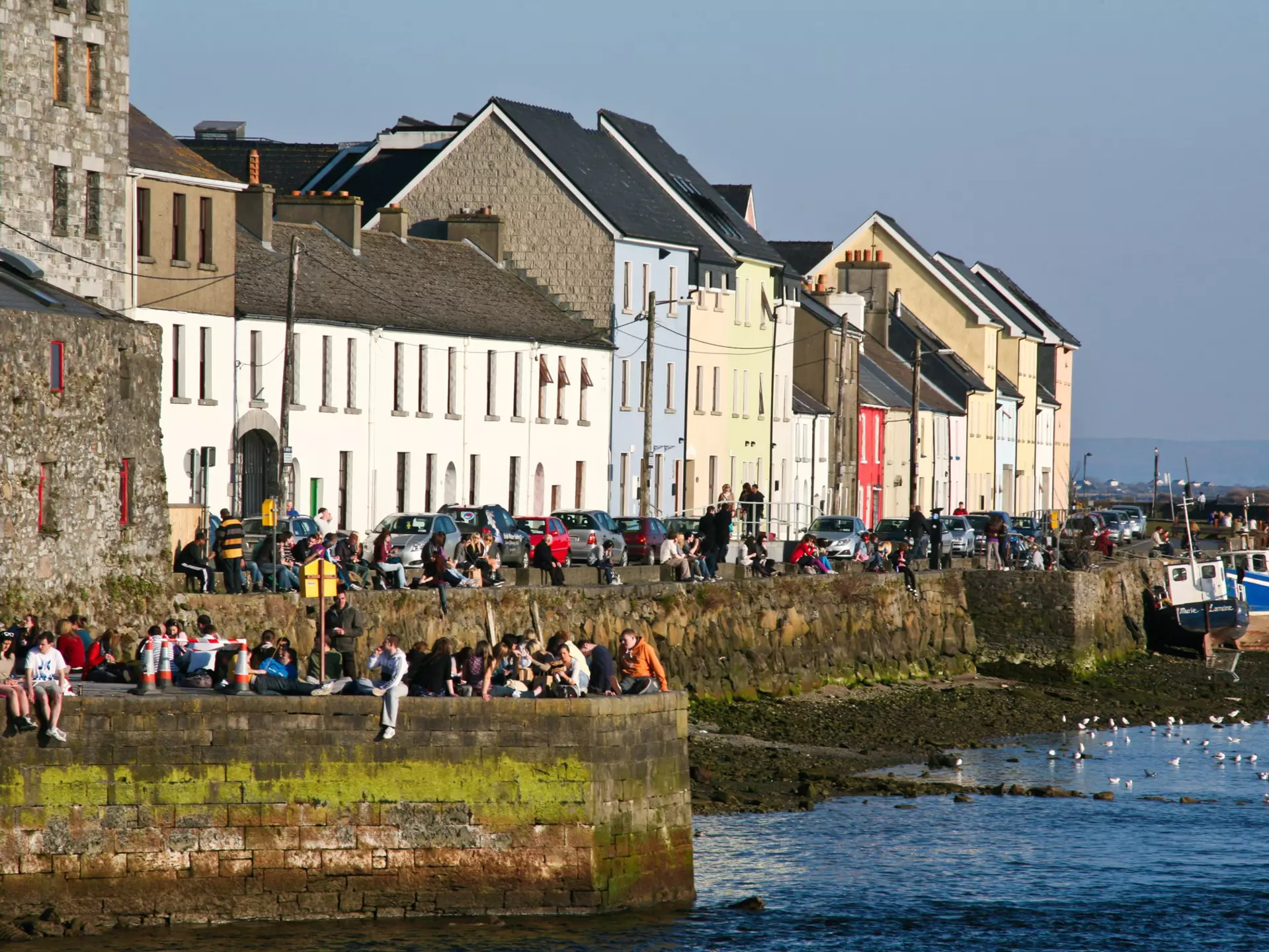 People sit on a sea wall in sunshine, with low buildings behind them.