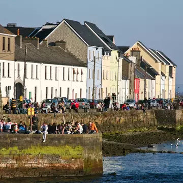 People sit on a sea wall in sunshine, with low buildings behind them.