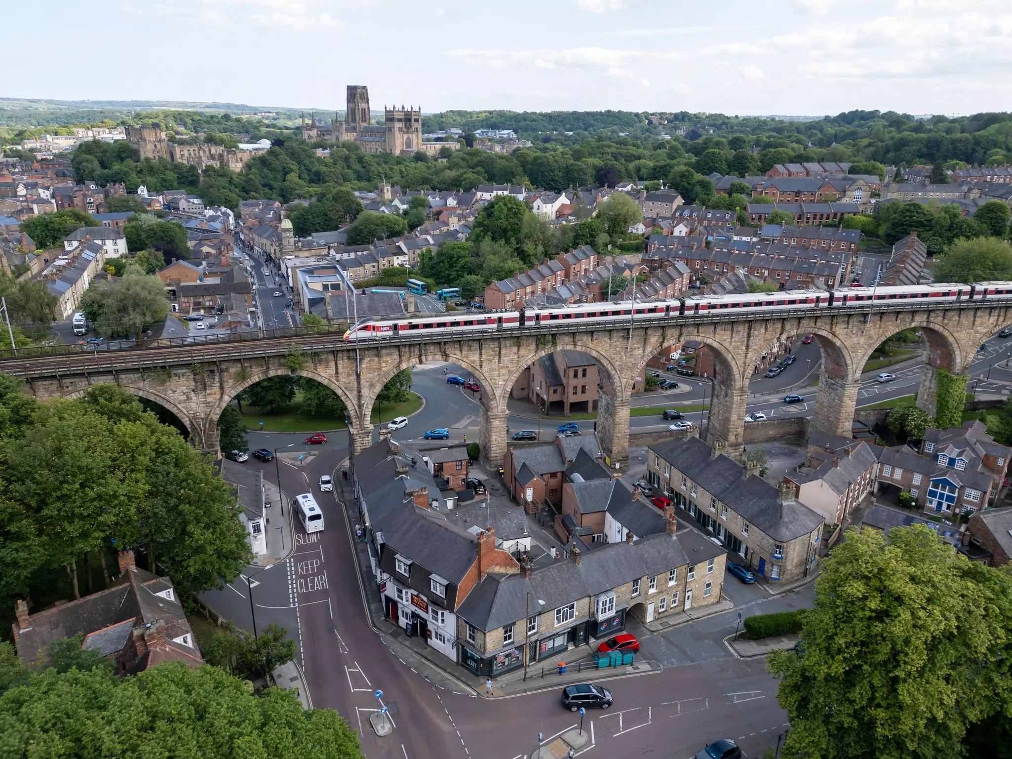 A train crosses a multi-arched viaduct in the center of a city. A large cathedral stands tall in the distance.