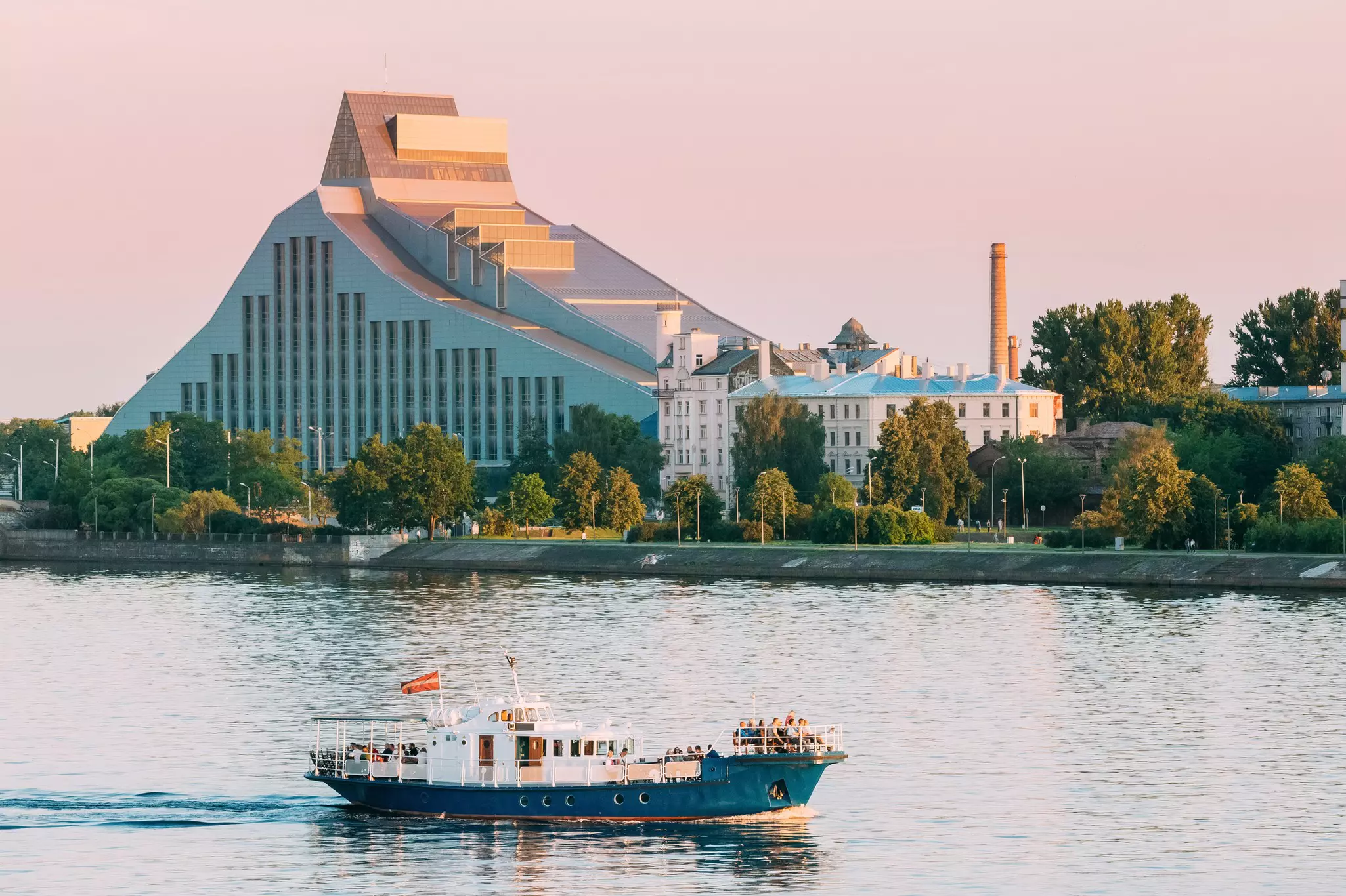 A passenger boat floats along a river in front of a large modern building with a sloped roof.