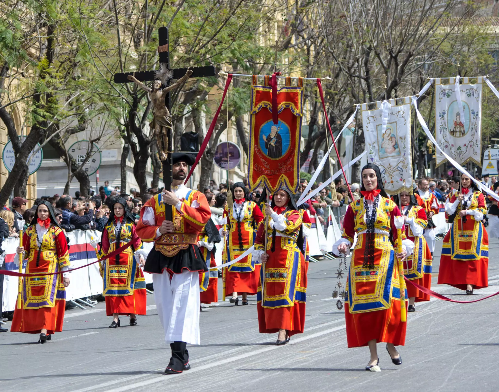 Local people in traditional Sardinian costumes walk along a street holding flags during a festival parade.