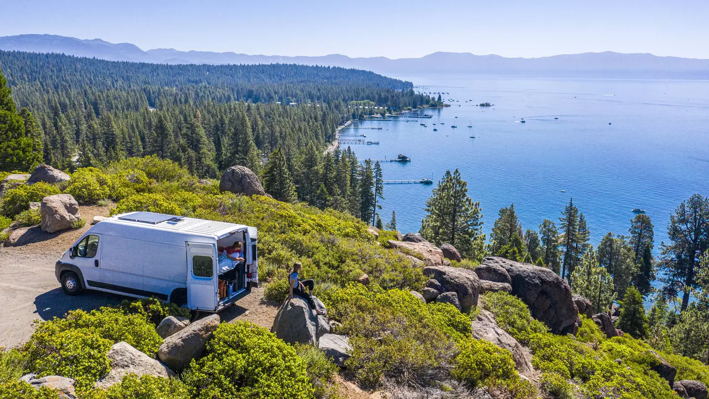 Aerial shot of a campervan at a viewpoint with two people sat overlooking a lake