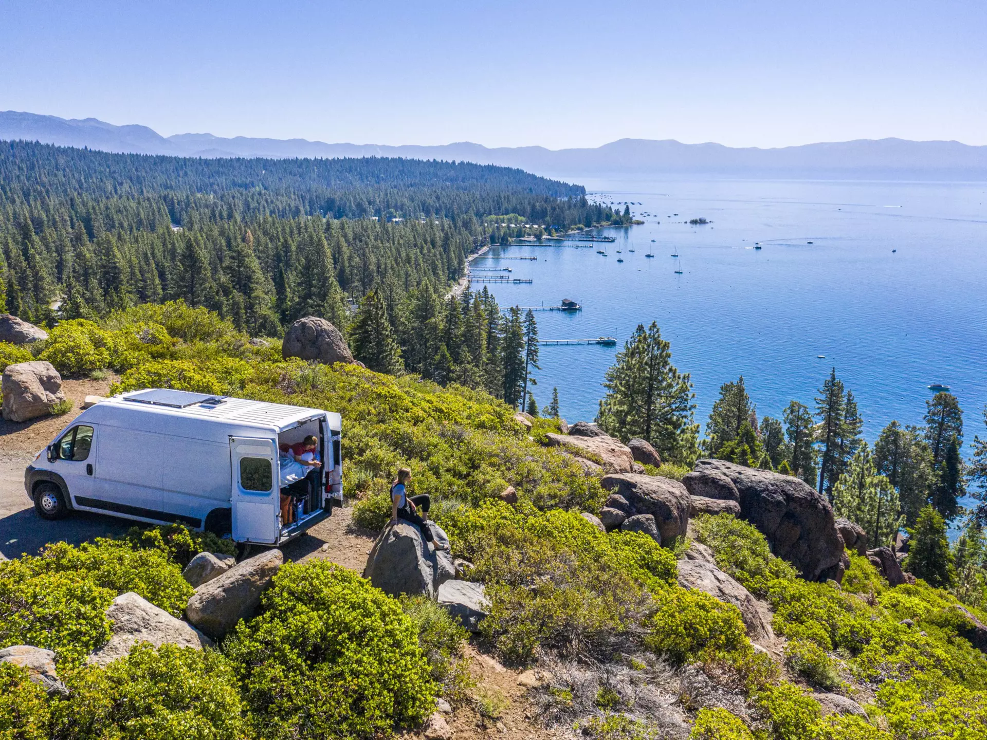 Aerial shot of a campervan at a viewpoint with two people sat overlooking a lake