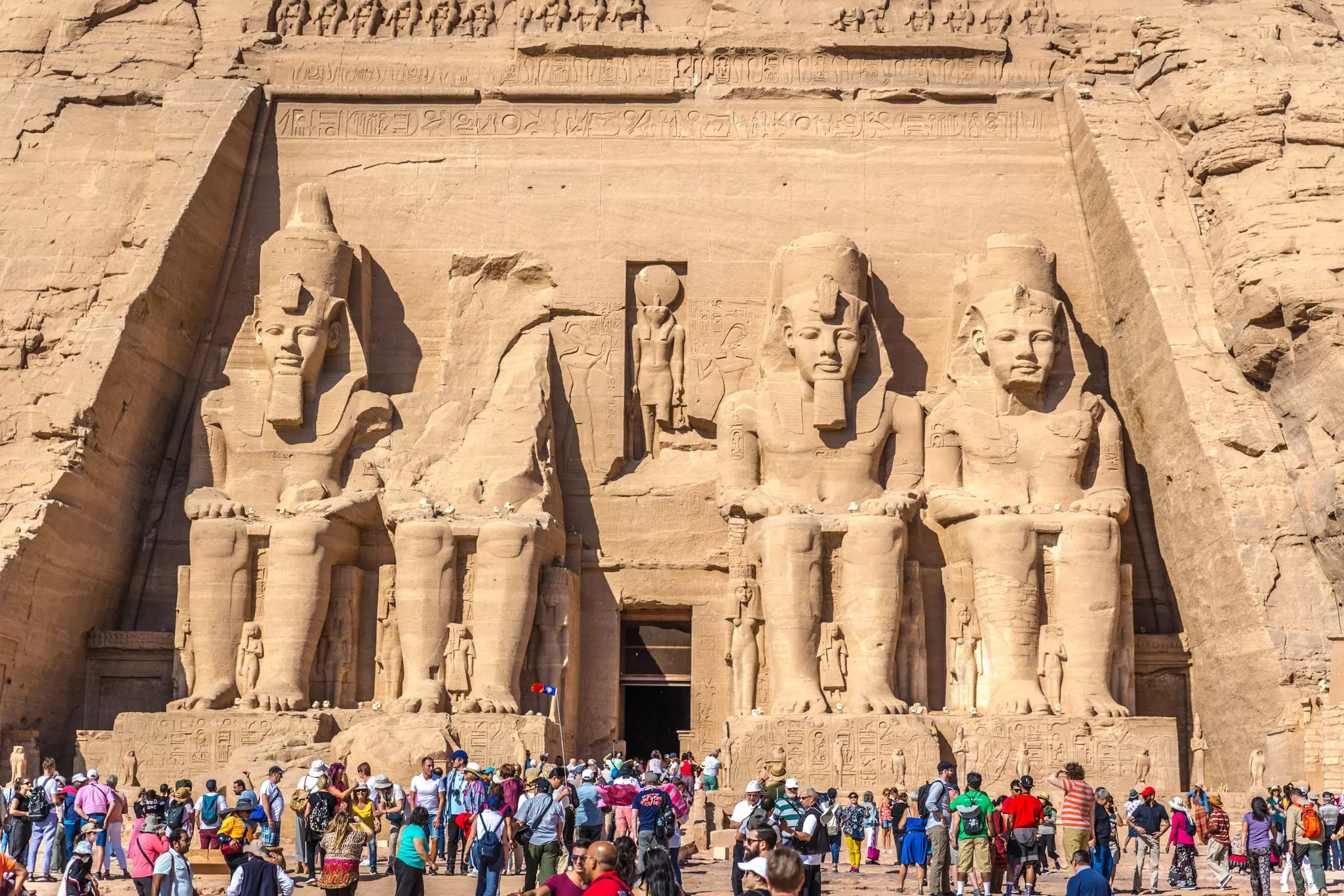 Tourists stand in front of a temple dominated by four huge sculptures of ancient Egyptian figures. The second seated figure is in ruins and no longer has a torso or head.
