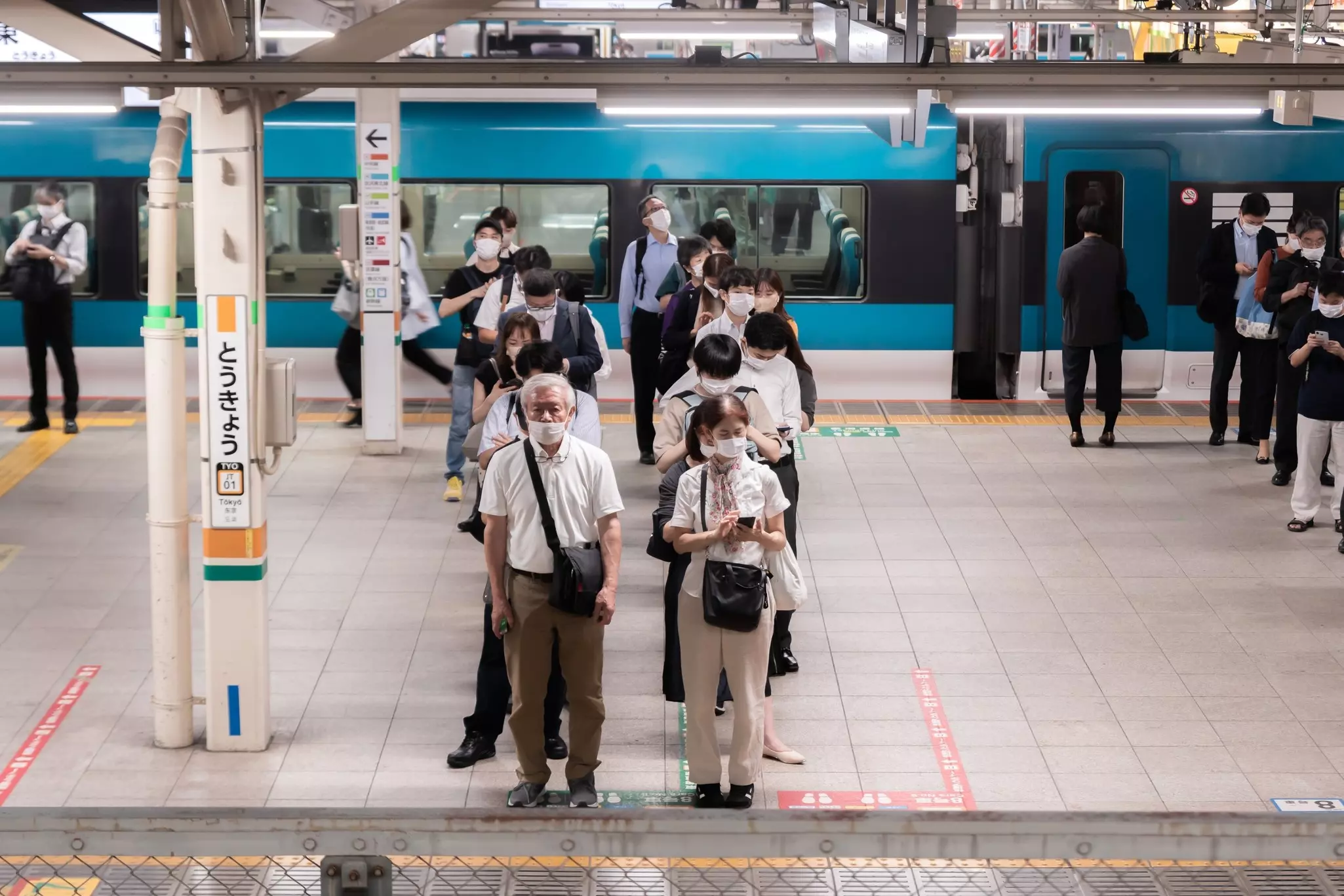 People wearing masks stand in a queue on a platform at a train station.