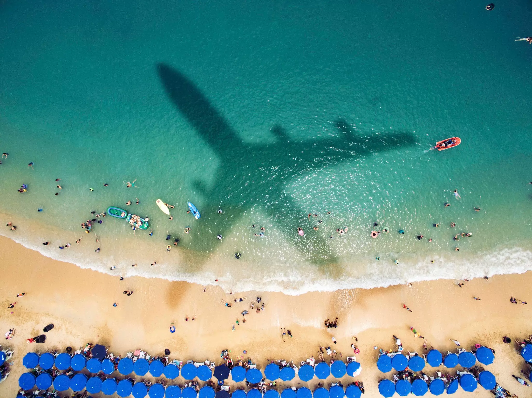 Aerial view of a crowded beach with blue umbrellas and swimmers as an airplane's shadow passes over the water