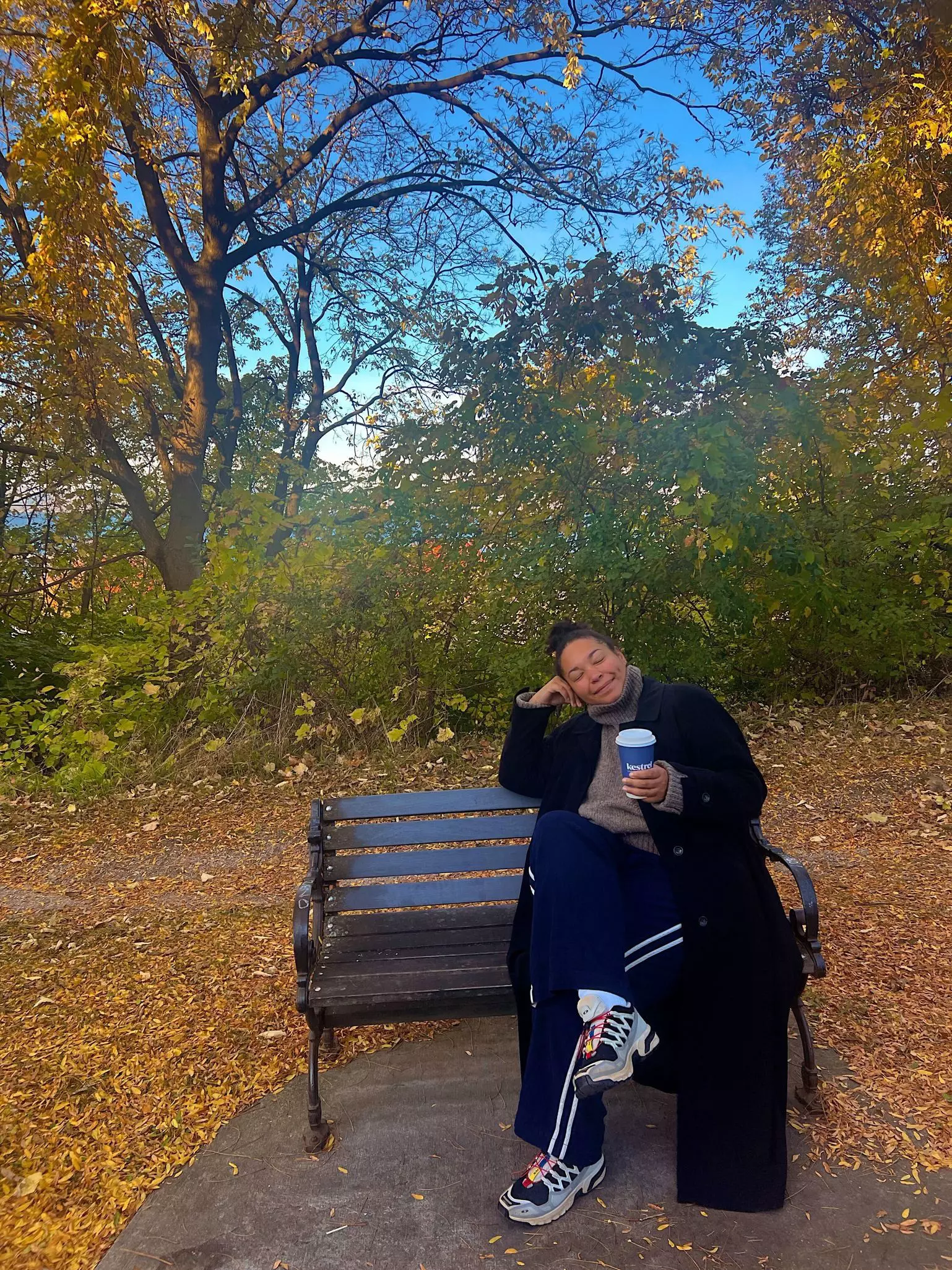 A woman wearing a sweater and long coat and holding a paper cup of coffee sits on a bench surrounded by golden leaves.