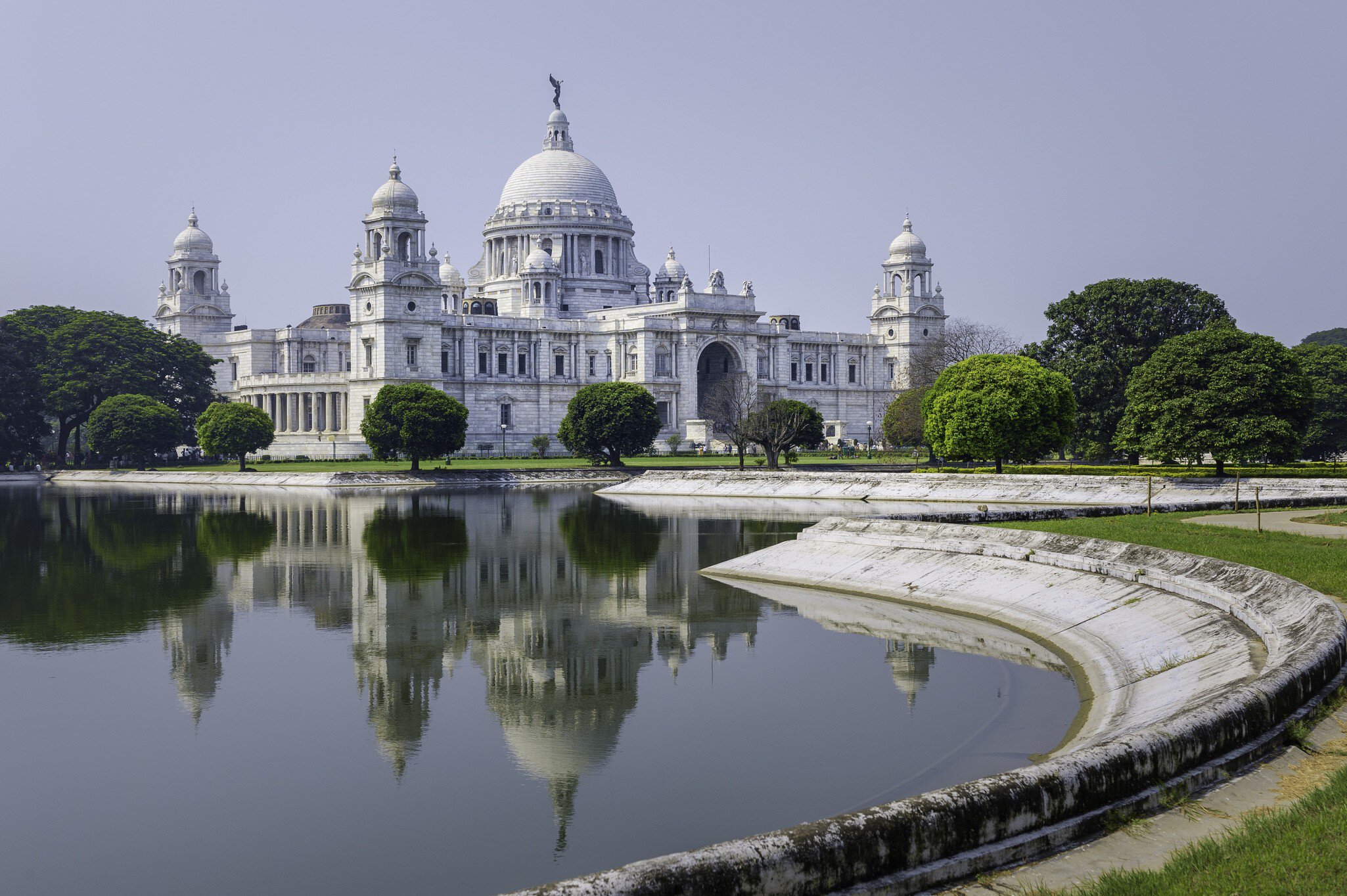Facade of The Victoria Memorial, a white marble monument in memory of Queen Victoria, on a bright sunny morning with blue sky with reflections of the memorial in the water feature to the front of the monument. Kolkata, India.