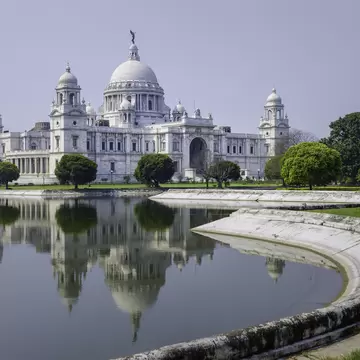 Facade of The Victoria Memorial, a white marble monument in memory of Queen Victoria, on a bright sunny morning with blue sky with reflections of the memorial in the water feature to the front of the monument. Kolkata, India.
