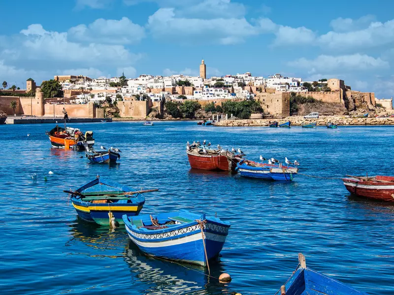 Boats in blue water; a fort is on the shore and a low-rise city behind it.