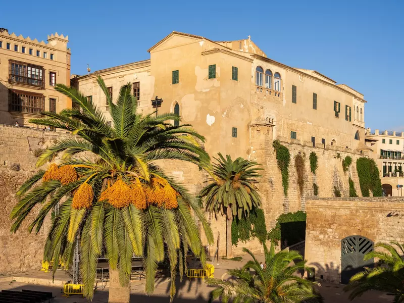 A honey-colored medieval palace framed by palm trees overlooks a courtyard.