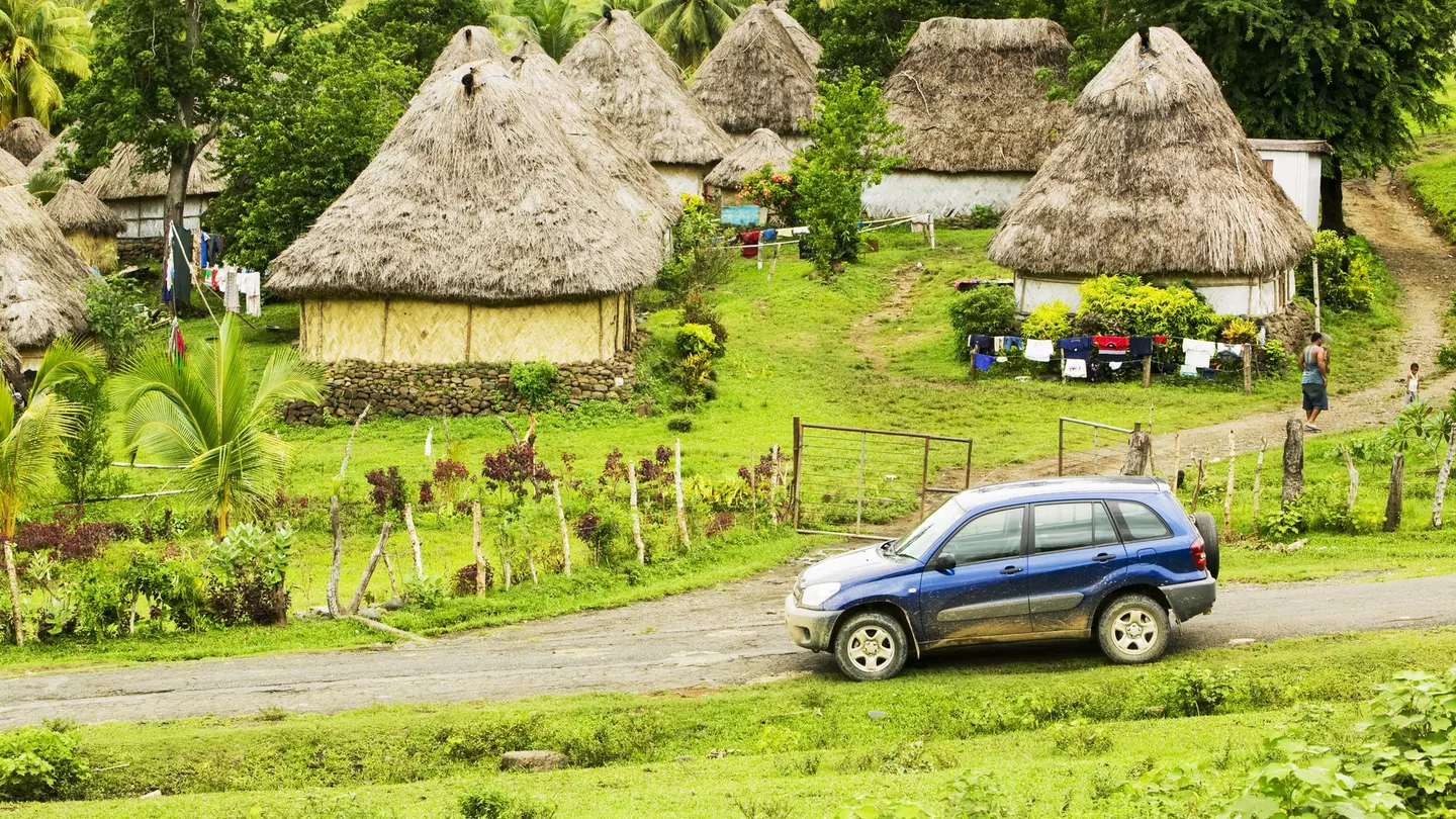A four-wheel drive car on a dirt track at the edge of a village of thatched huts.