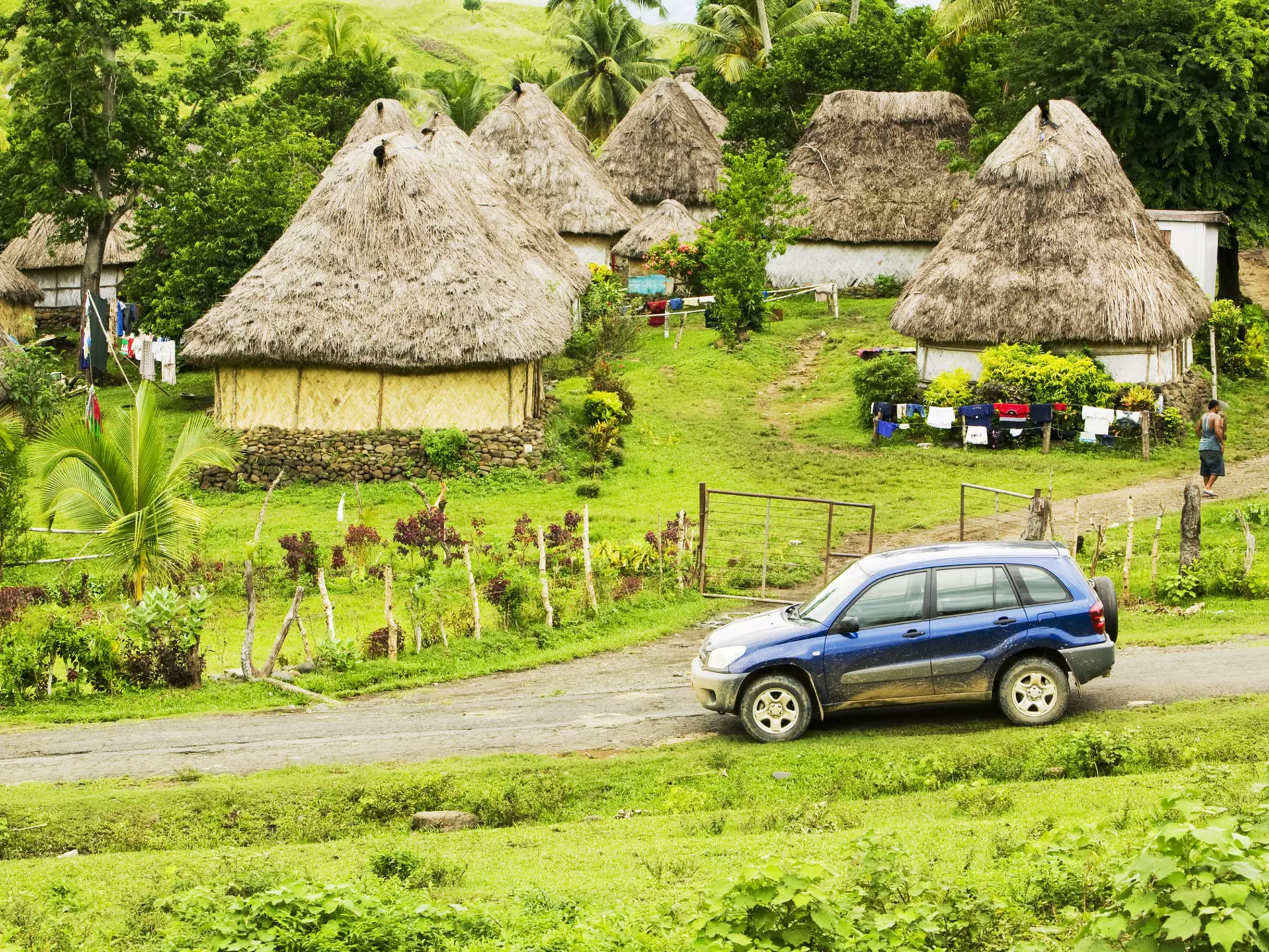 A four-wheel drive car on a dirt track at the edge of a village of thatched huts.