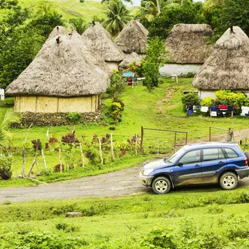 A four-wheel drive car on a dirt track at the edge of a village of thatched huts.
