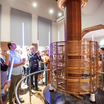 A tall copper Tesla coil discharging a violet arc to a ceiling electrode, watched by visitors holding fluorescent tubes.