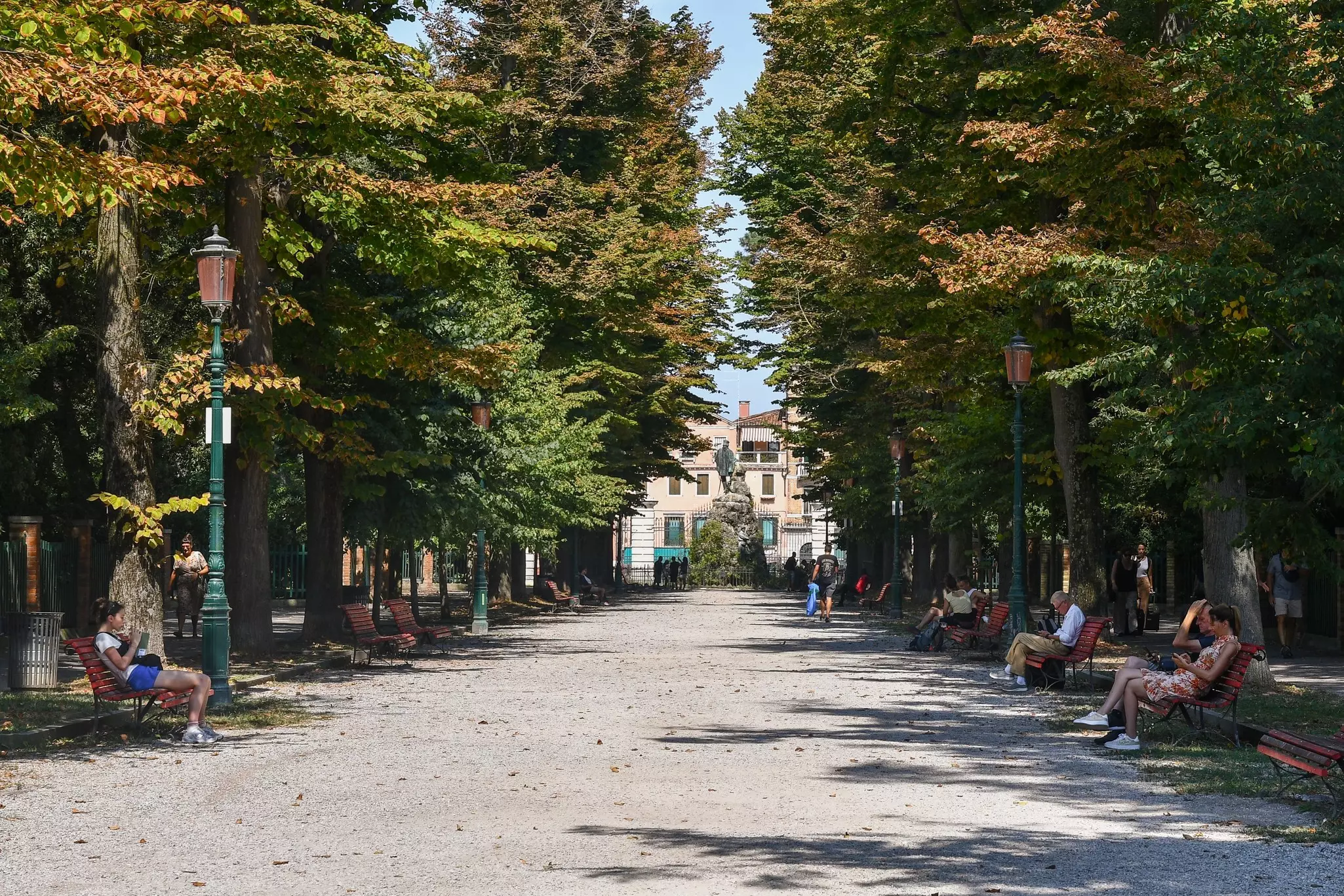 A gravel alley in a city park is pictured. People sit on benches in the shade of trees on either side of the alley, and a bronze sculpture is seen at the alley’s end.