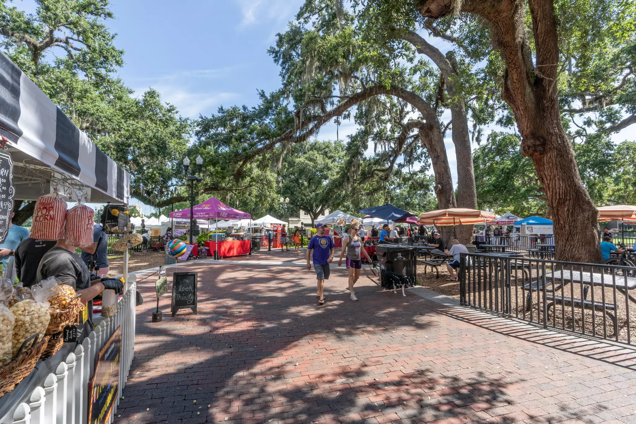 A cluster of booths with colorful umbrellas along a brick walkway under trees with moss hanging from them