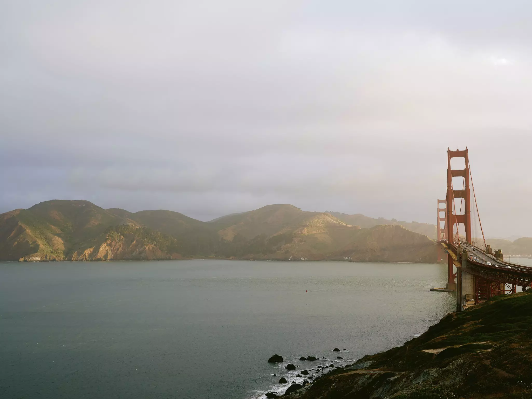 The orange arches of the Golden Gate Bridge stretch over the ocean with hills in the background and a thin layer of fog