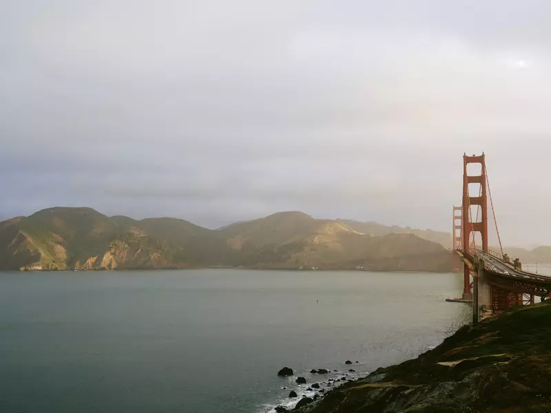 The orange arches of the Golden Gate Bridge stretch over the ocean with hills in the background and a thin layer of fog