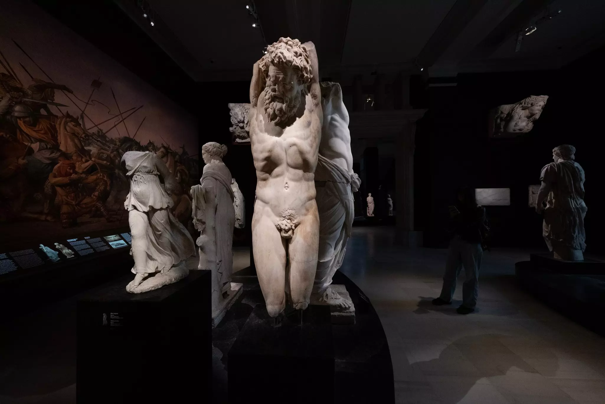 A sculpture in a darkened museum gallery depicts a bearded, nude figure raising his arms in fear.