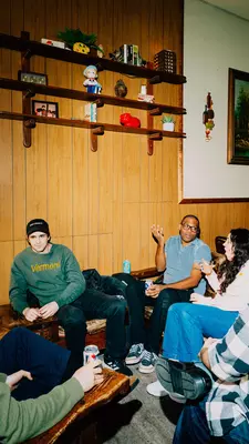 A man in a white t-shirt and a khaki jacket with a shot in one hand and a PBR in the other.