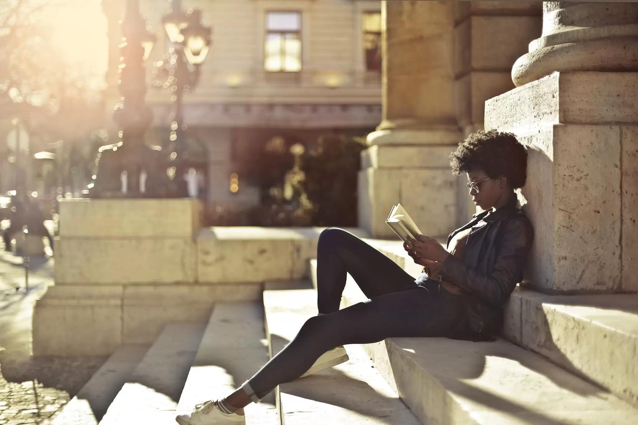 Woman in a leather jacket reads a book while seated on sunlit stone steps beside a classical column at golden hour.