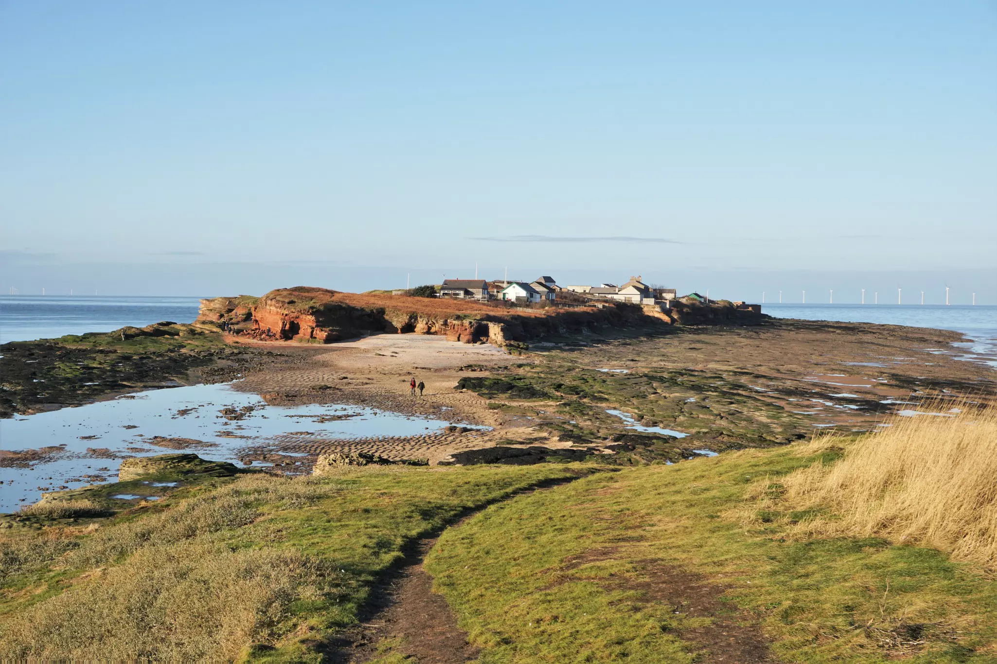 Looking towards Hilbre Island from Little Hilbre Island, located in the estuary of the River Dee. At low tide people can walk from West Kirby to the Hilbre Islands.  Two walkers are walking along the exposed river bed.